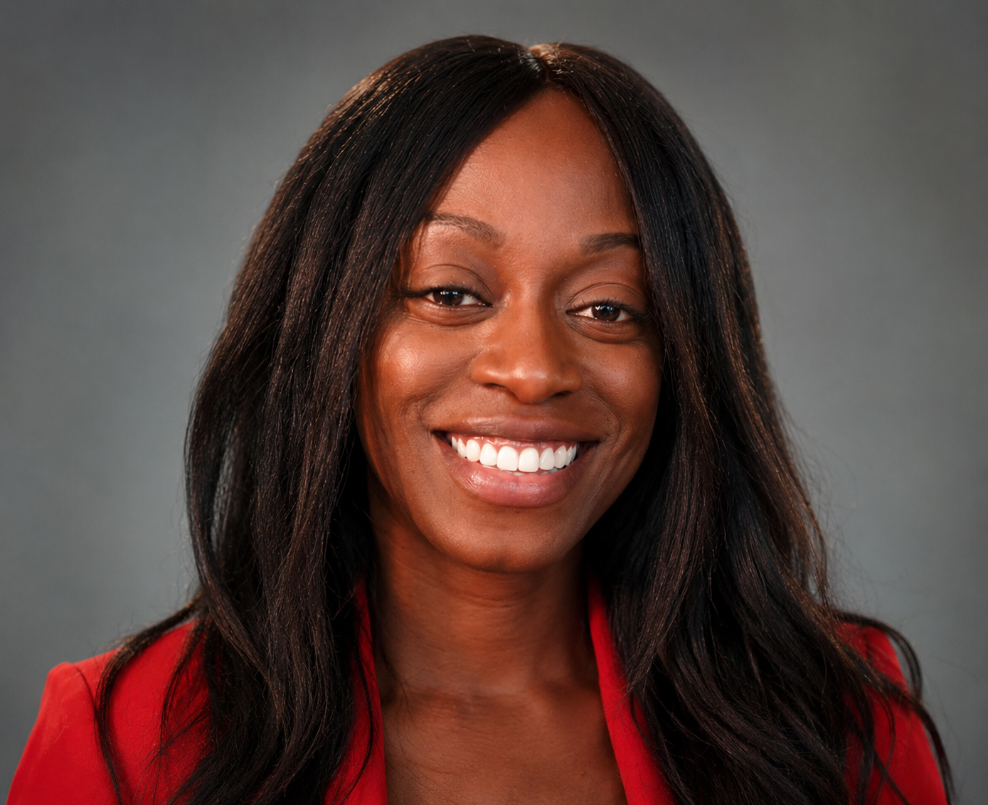 Portrait of DeLora Lambert, MFA — a Black woman in a red blazer with long dark hair, smiling warmly at the camera against a soft grey studio backdrop.